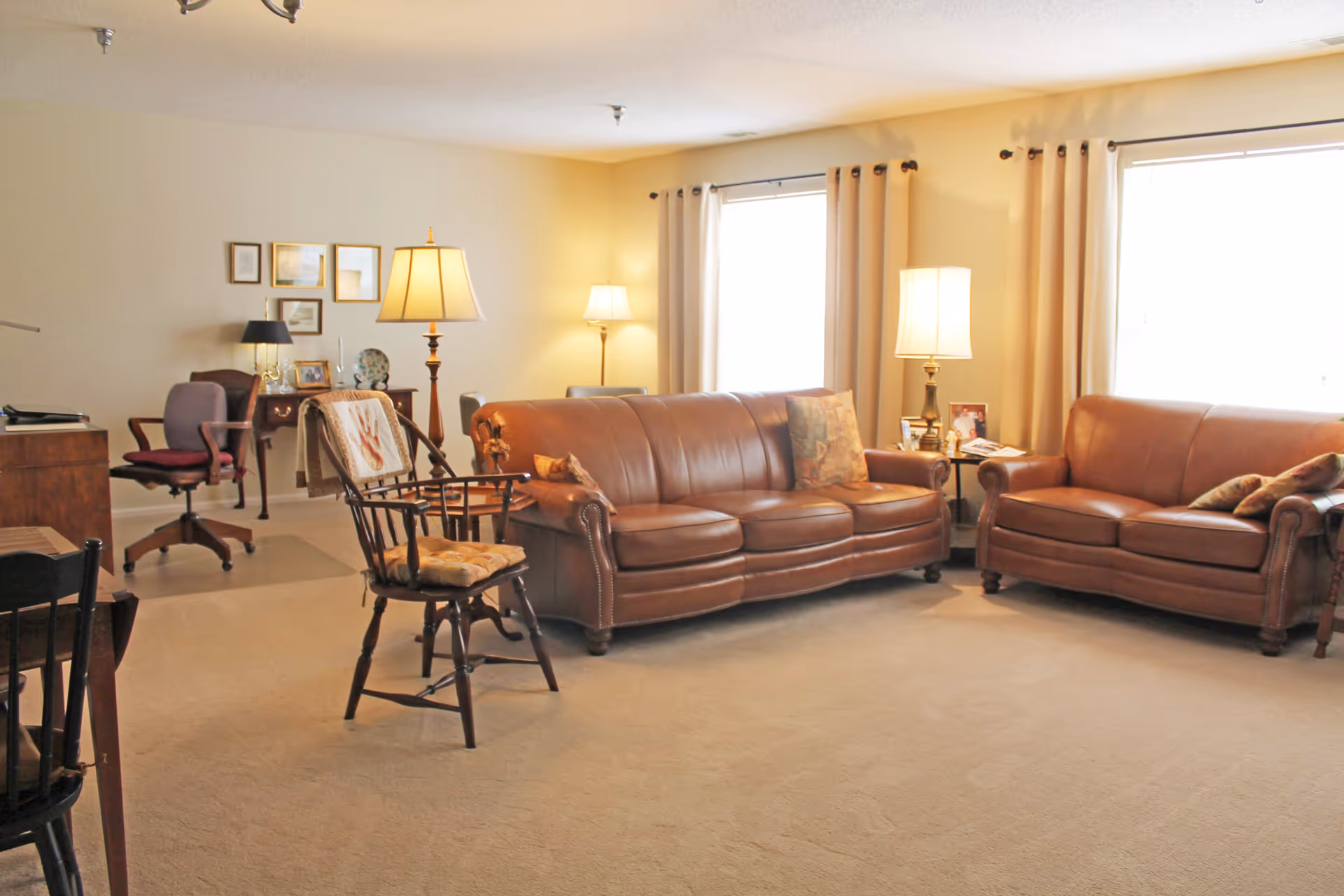 A cozy living room with two brown leather sofas, a wooden chair with a cushion, two side tables with lamps, and a desk with a chair in the background. The room is well-lit with natural light coming through two windows with beige curtains.