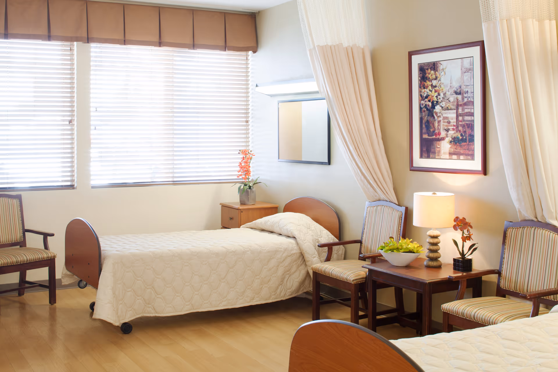 A bright senior living bedroom with two single beds, wooden chairs, a side table with a lamp and framed artwork on the wall.