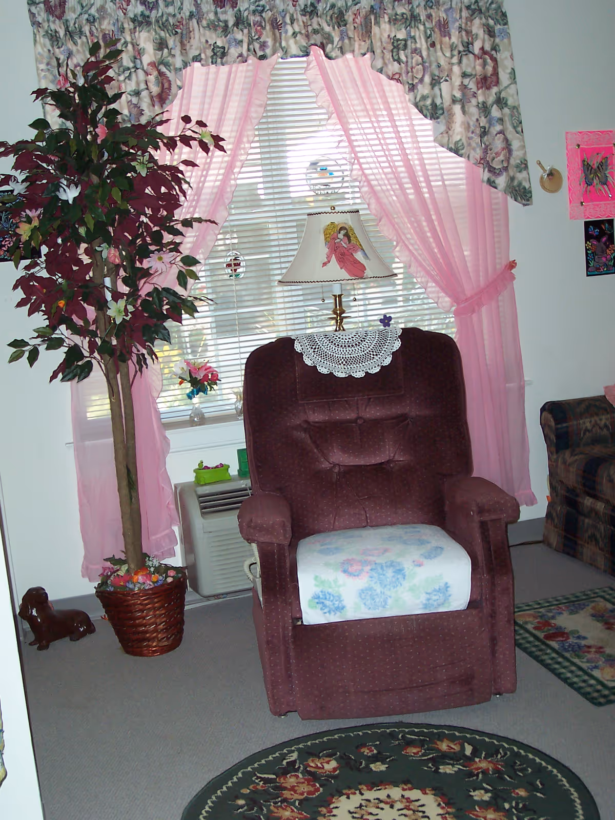 A cozy living room corner featuring a maroon recliner chair with a floral cushion and a white doily on the headrest. Behind the chair is a window with pink sheer curtains and floral valance. A decorative lamp with an angel design sits on the windowsill. To the left of the chair is a tall potted artificial tree, and to the right, part of a patterned sofa is visible. The floor has a floral round rug and a rectangular rug with a floral pattern.