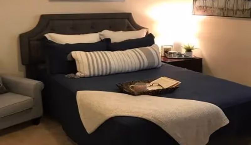 A neatly made bed with a dark upholstered headboard, dark blue bedding, multiple pillows including a long striped one, and a light-colored throw blanket at the foot. A tray with a cup, glasses, and an open book rests on the bed. To the left is a gray armchair, and to the right is a nightstand with a lamp, a small plant, and framed pictures. The room has warm lighting and a piece of wall art above the nightstand.
