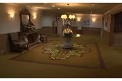 Dimly lit interior lobby with patterned carpet, a central floral arrangement on a pedestal table, and seating along the walls.