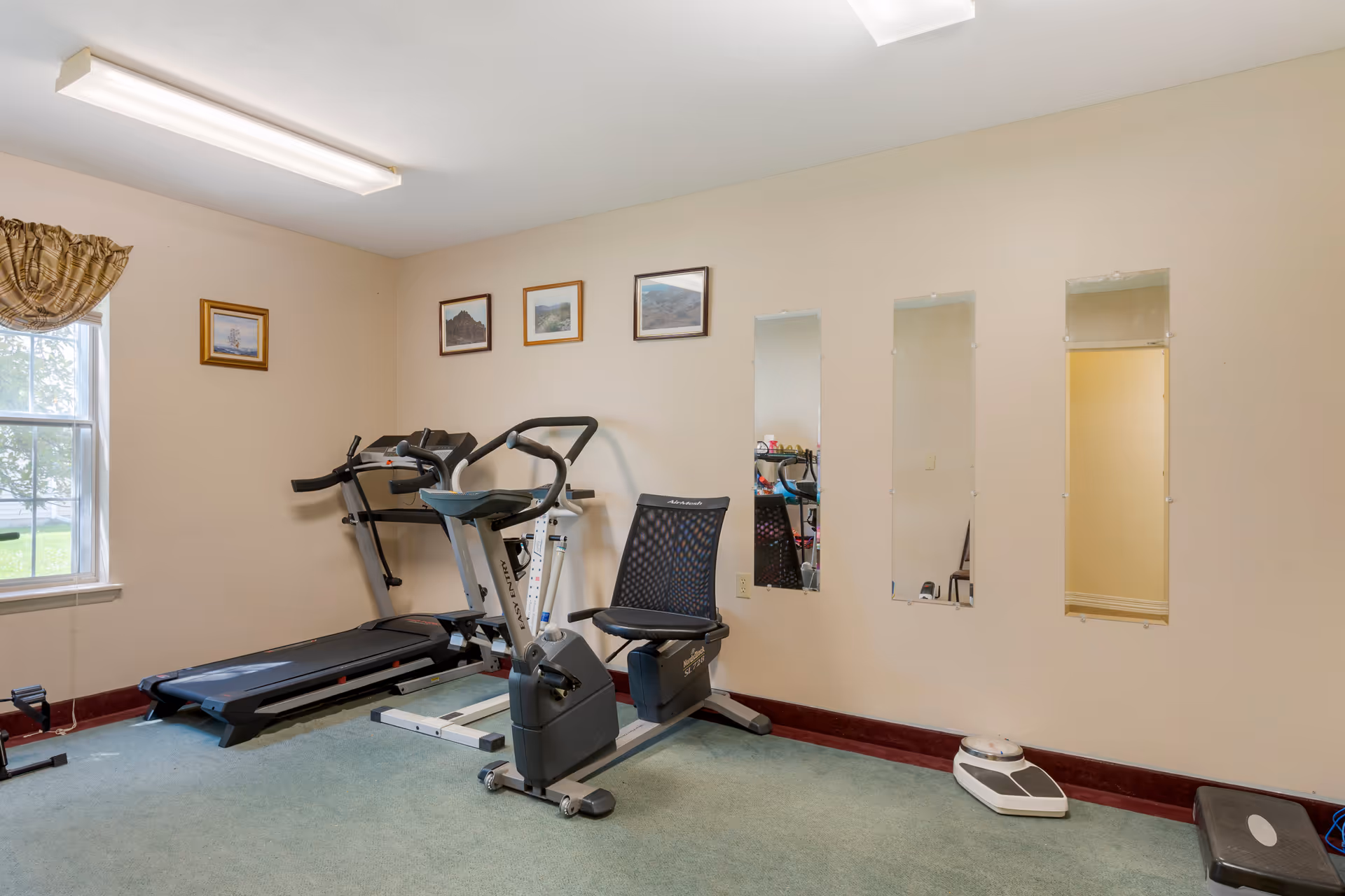 A small exercise room with a treadmill, a stationary recumbent bike, three vertical mirrors on the wall, a window with a curtain, framed pictures on the wall, a scale on the floor, and a step platform.