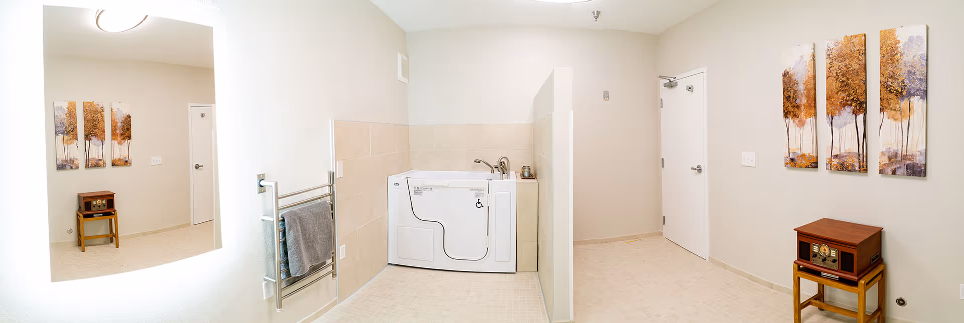 A spacious bathroom with a walk-in bathtub designed for accessibility. The walls are painted light beige with beige tiles halfway up. There is a large illuminated mirror on the left wall, a towel rack with a gray towel beneath it, and a small wooden table with a vintage-style radio and a three-panel artwork of trees on the right wall. The floor is tiled in a light color, and there is a white door on the right side of the room.