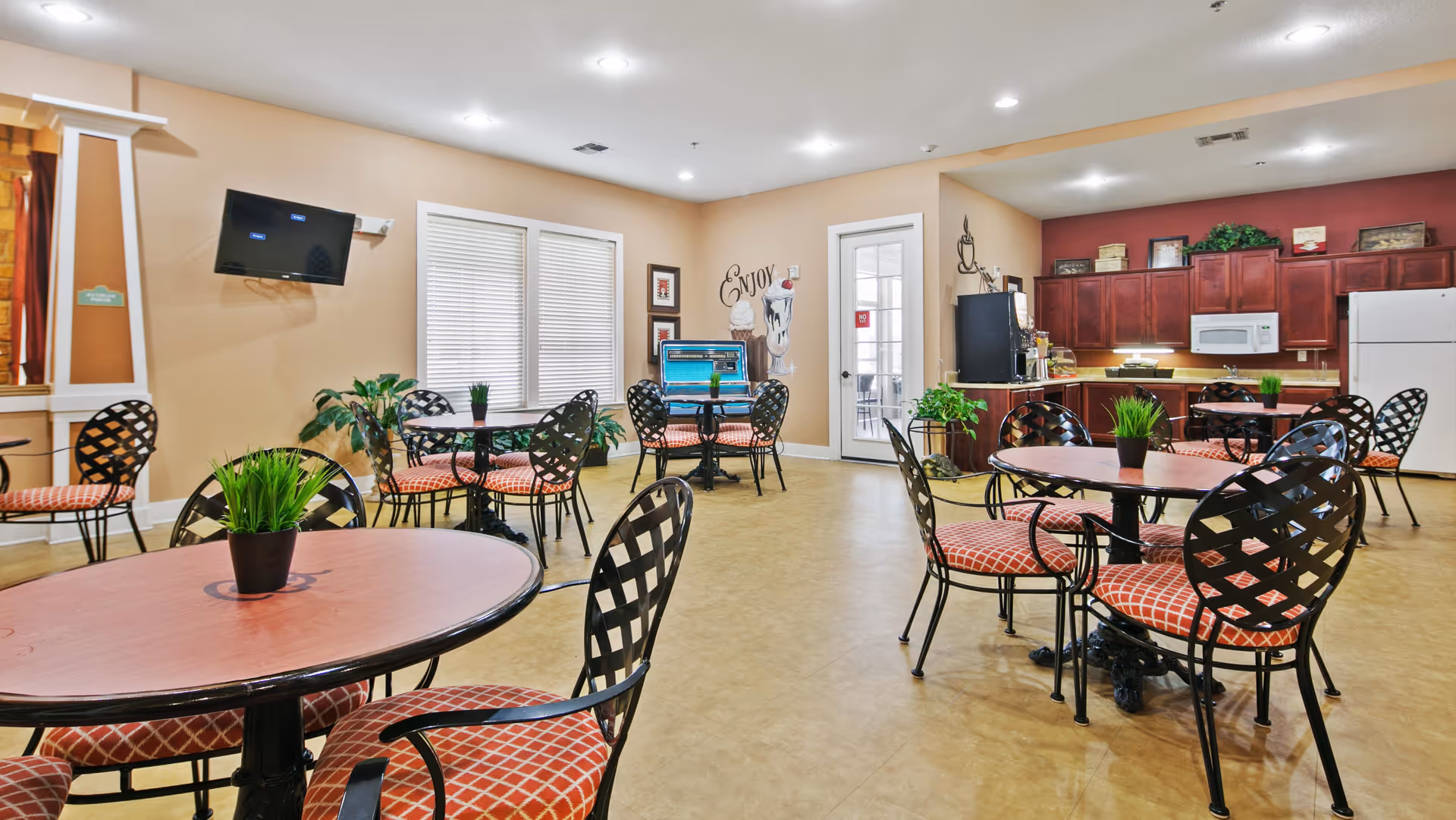 A bright and spacious dining area with several round tables and black metal chairs with red patterned cushions. The room features beige walls, a mounted flat-screen TV, large windows with blinds, and a kitchen area with dark wood cabinets, a white refrigerator, and a microwave. There are small green plants on the tables and decorative wall art including the word 'Enjoy'.