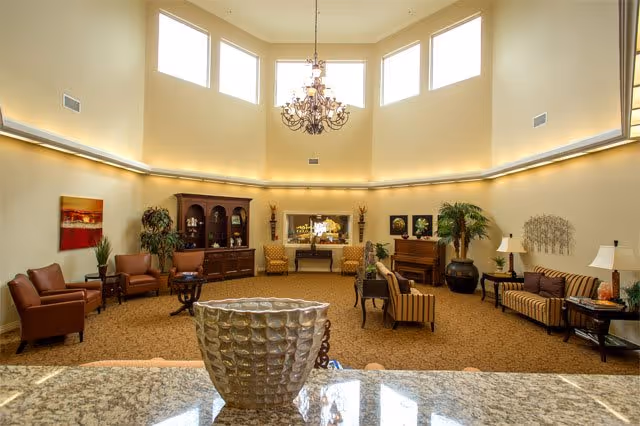 Spacious senior living common room with high windows, a chandelier, fireplace, and multiple seating areas.