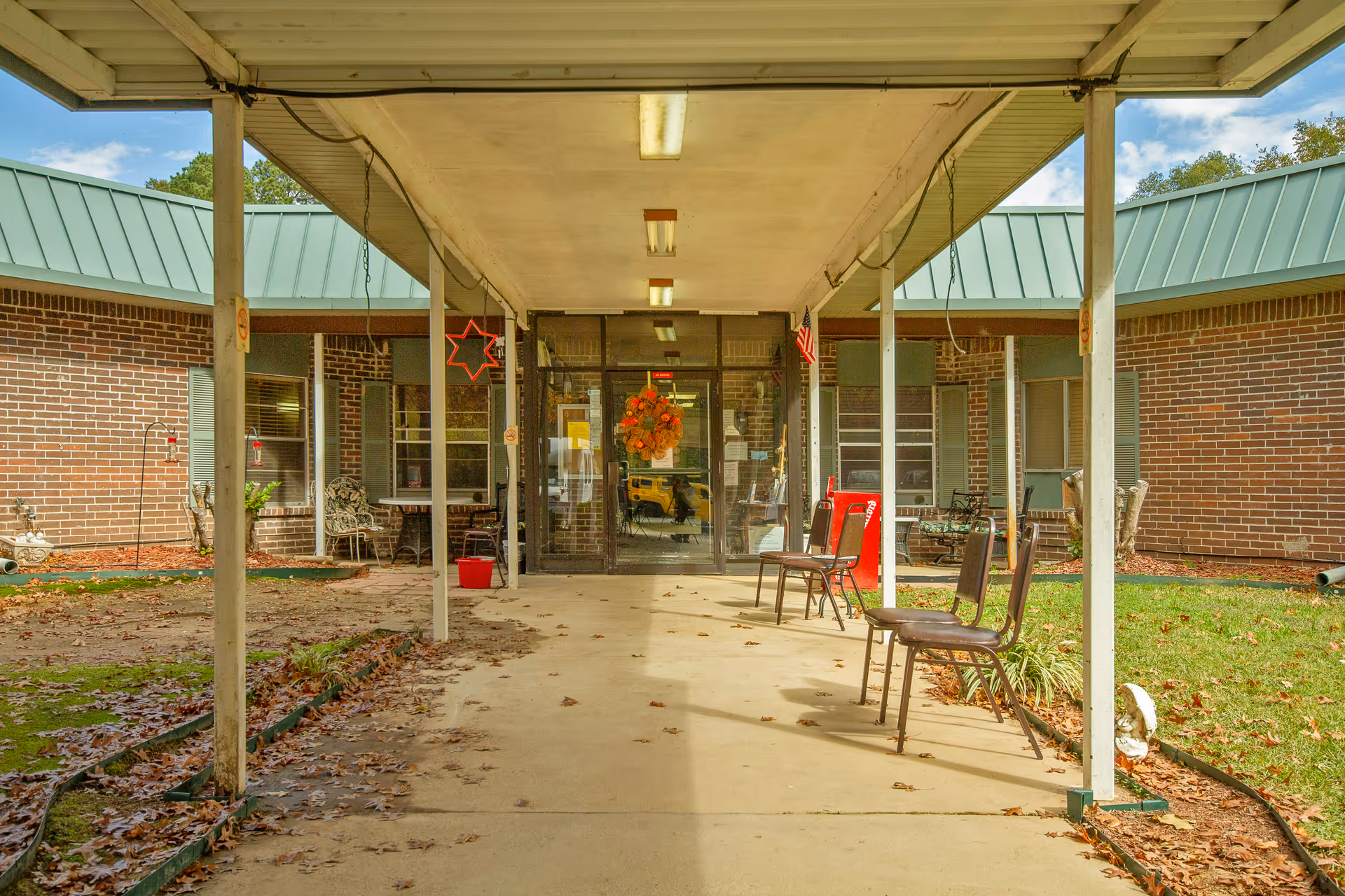 Covered walkway leading to the entrance of a brick building with green metal roofing. There are several chairs lined up along the right side of the walkway, a red Coca-Cola vending machine near the entrance, and autumn leaves scattered on the ground. The entrance door has a fall-themed wreath and an American flag is hanging nearby.