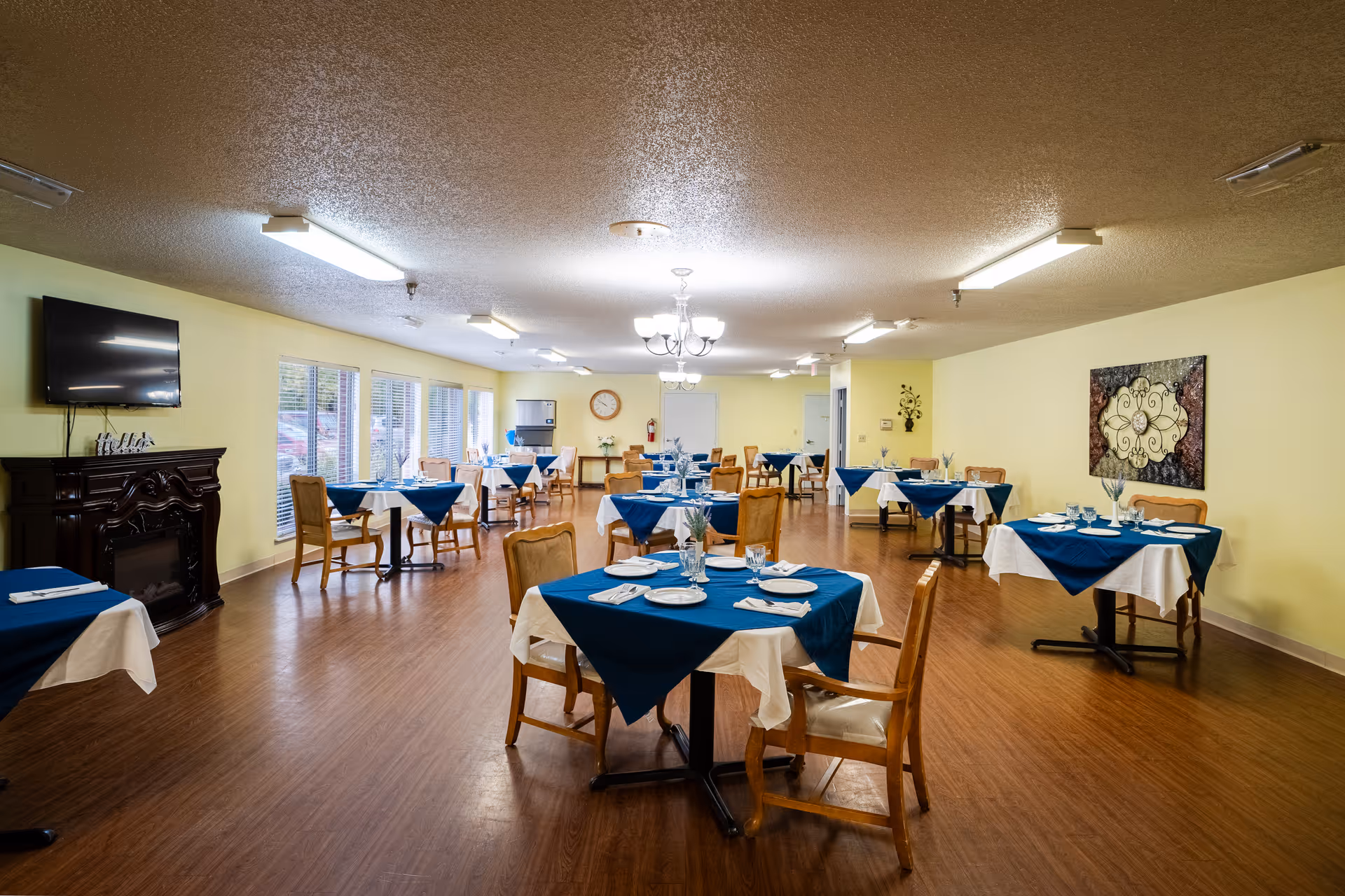 A bright dining room with multiple tables set with blue tablecloths and wooden chairs.