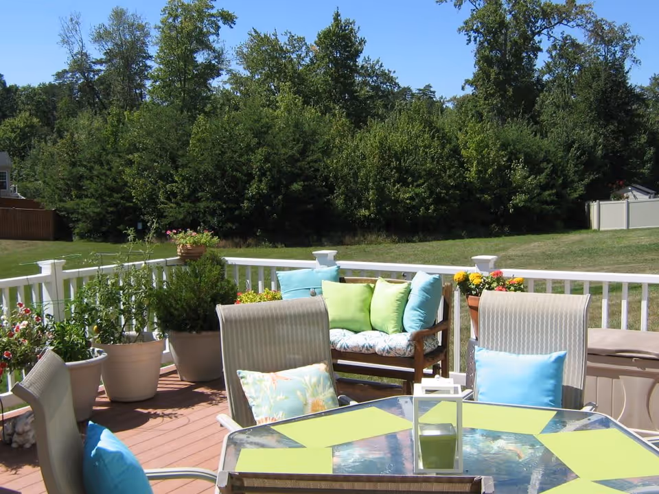 Outdoor patio area with a glass table and several chairs, some with blue and green cushions. There is a wooden bench with green and blue pillows, and various potted plants and flowers around the patio. In the background, there is a grassy yard and dense green trees under a clear blue sky.