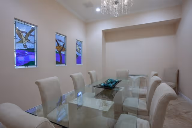 A dining room with a glass table surrounded by eight beige upholstered chairs. The room features three narrow stained glass windows with bird designs on one wall and a crystal chandelier hanging from the ceiling.