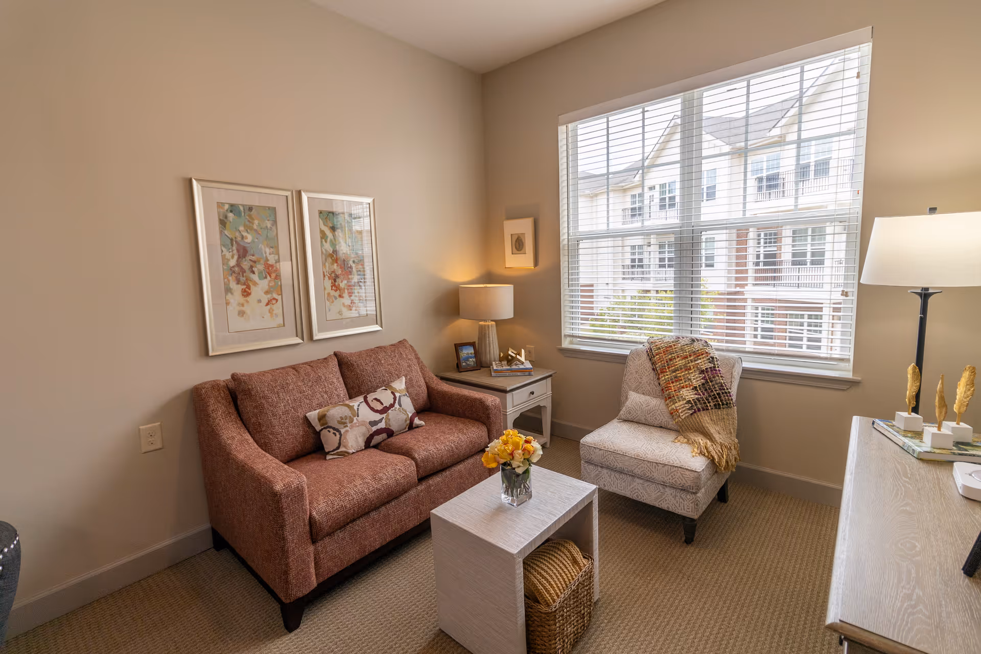 A cozy living room with a red sofa, a patterned armchair with a colorful throw blanket, a small white coffee table with a vase of flowers, a side table with a lamp and framed photo, two framed abstract paintings on the wall, and a large window with blinds letting in natural light.