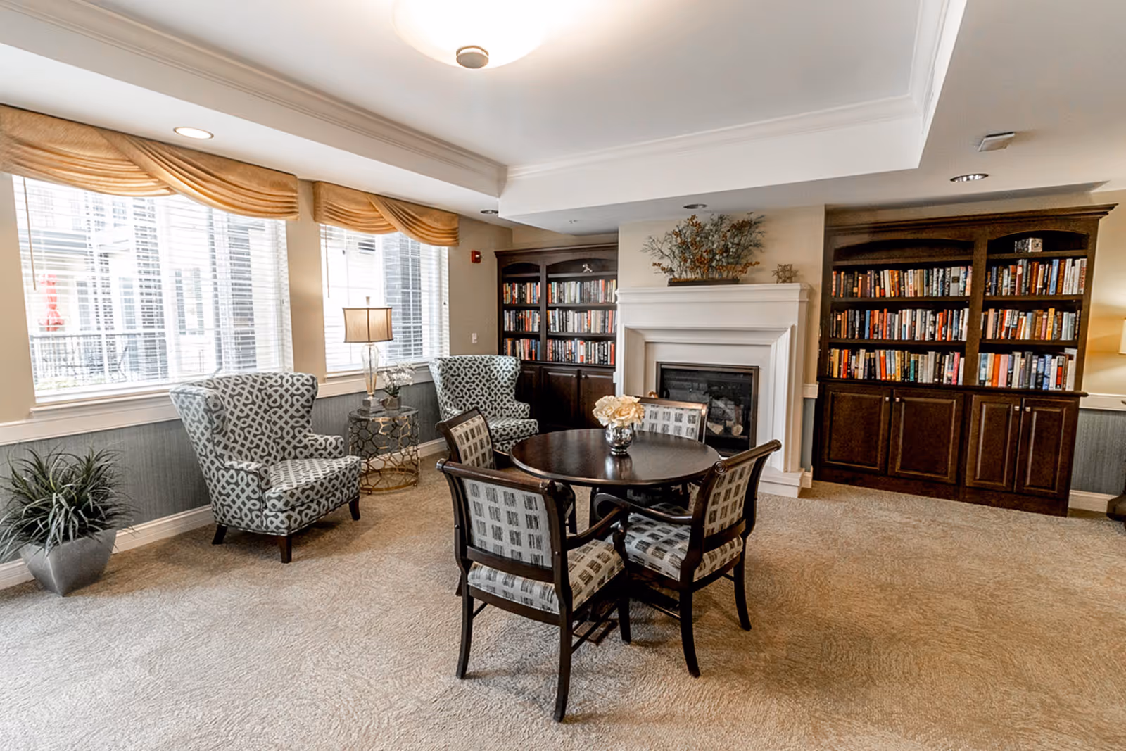 Cozy communal sitting room with a round table and chairs, patterned armchairs by bright windows, a fireplace and built-in bookshelves.