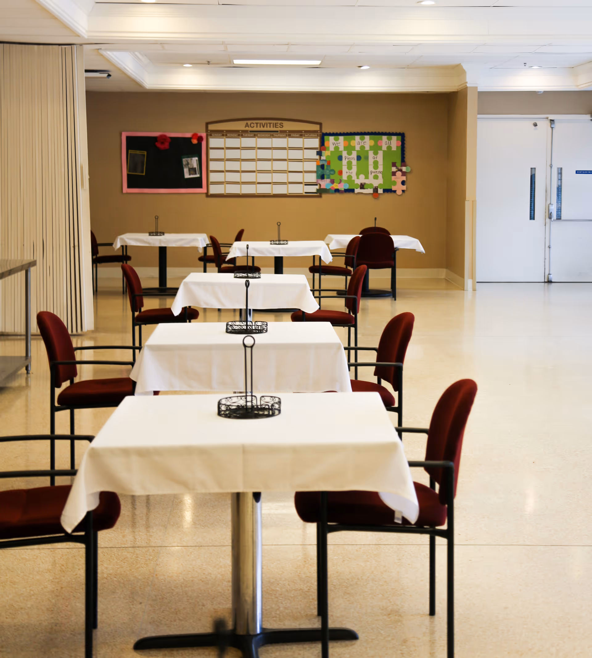 Empty dining room in a senior facility with rows of small tables draped in white cloths and red upholstered chairs.