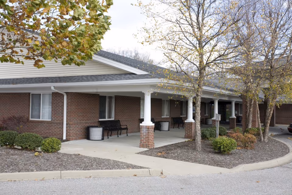 Covered brick entrance of a senior living facility with white columns, benches, and trees in front.
