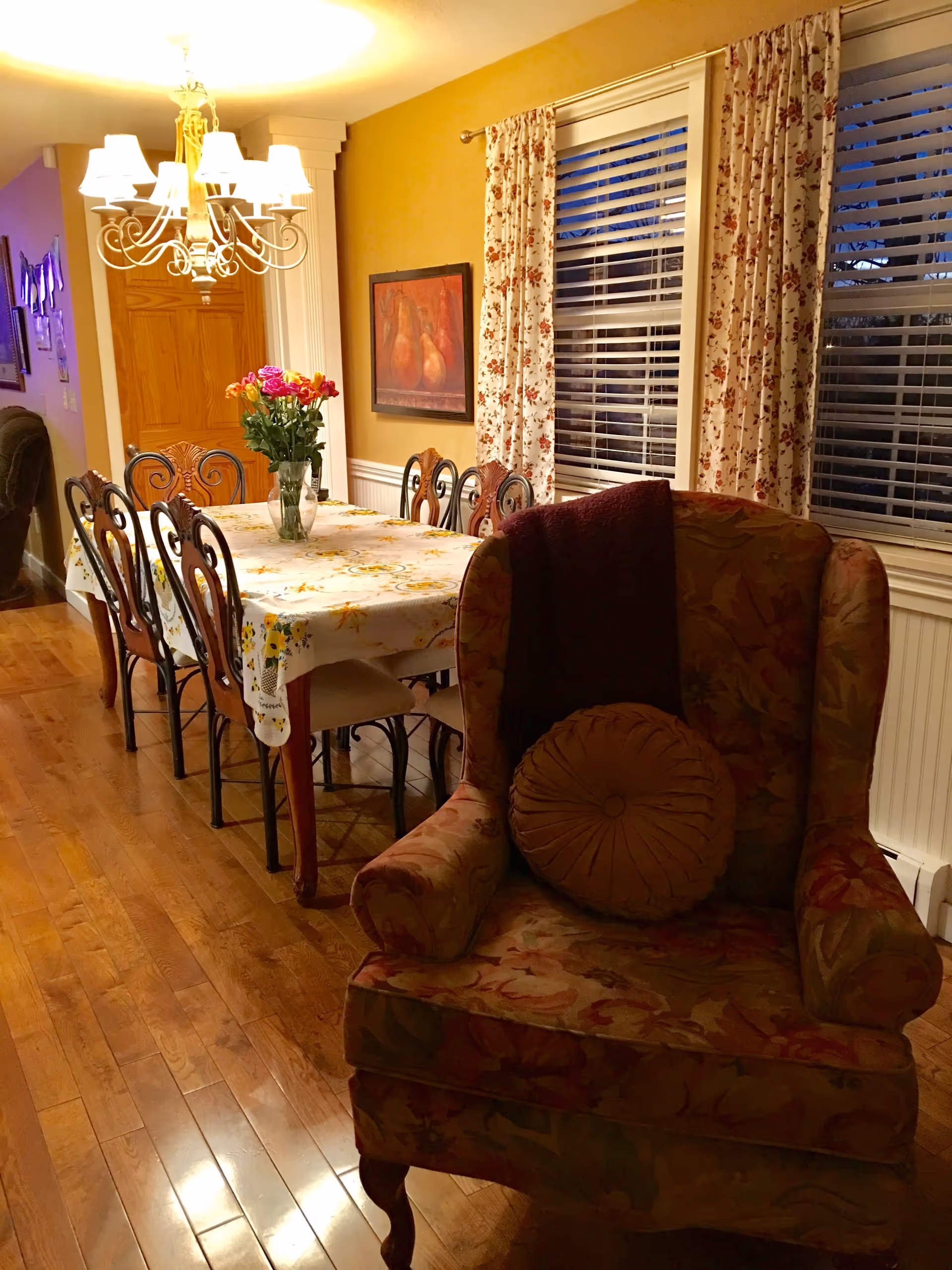 A warmly lit dining room with a wooden floor, a long dining table covered with a floral tablecloth, and eight chairs. A vase with colorful flowers is placed on the table. To the right, there is a floral upholstered armchair with a round cushion and a dark throw draped over the back. The wall behind the dining table is painted yellow and decorated with a framed painting of pears. Two windows with floral curtains and white blinds are visible on the right side.