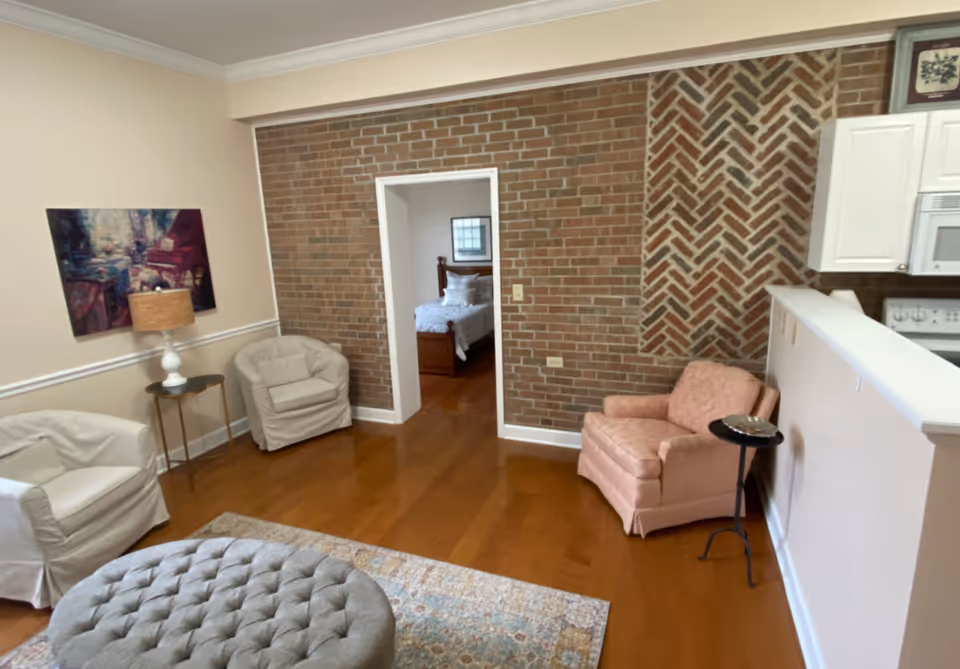 A cozy living room with wooden floors, a patterned area rug, two white armchairs, a pink armchair, a round tufted ottoman, and a small side table with a lamp. The room features a brick accent wall with a doorway leading to a bedroom with a bed and window. Part of a white kitchen counter and cabinets are visible on the right.
