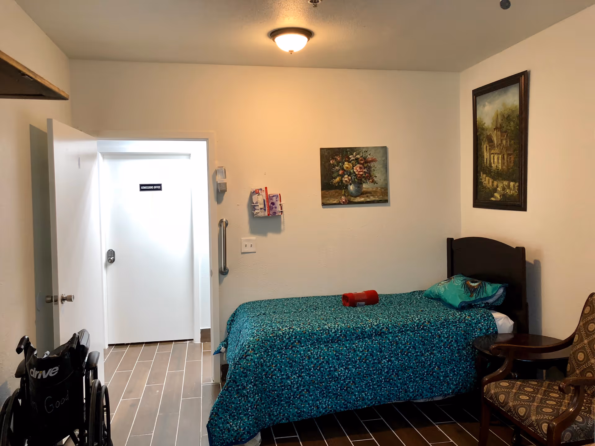 A small bedroom in an assisted living facility with a single bed covered in a teal patterned blanket, a pillow, and a red item on the bed. There is a wooden chair with patterned upholstery next to a small round table. Two framed paintings hang on the walls. A wheelchair is positioned near the open door, which leads to a hallway with a door labeled 'Admissions Office'. The floor is tiled with brown rectangular tiles.