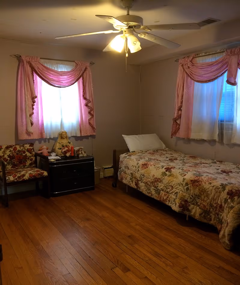 A bedroom in a senior living facility with floral bedspread, pink curtains, and a ceiling fan.