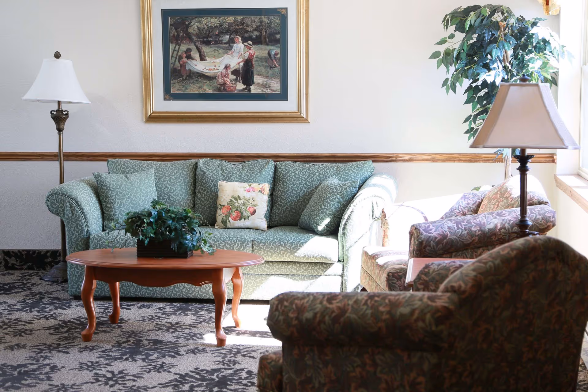 Sunlit sitting area with a patterned sofa, armchairs, a wooden coffee table with a plant, lamps, framed artwork, and a potted tree.