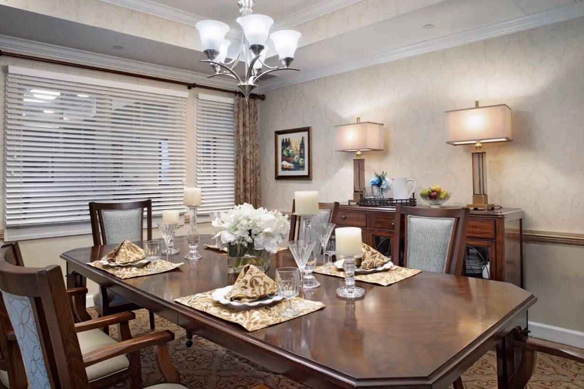 A formal dining room with a polished wooden table set for four, featuring patterned napkins, glassware, and candles. The room has a large window with white blinds, floral curtains, a chandelier overhead, and a wooden sideboard with two table lamps, a pitcher, a bowl of fruit, and a small floral arrangement. A framed painting hangs on the wallpapered wall.