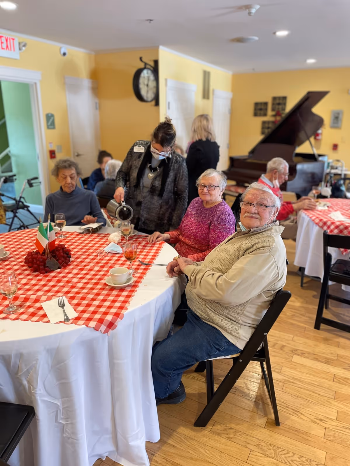 A group of elderly people sitting around a table with a red and white checkered tablecloth in a senior living facility dining room. A staff member wearing a mask is pouring a drink for one of the residents. There is a grand piano in the background and yellow walls with white doors.