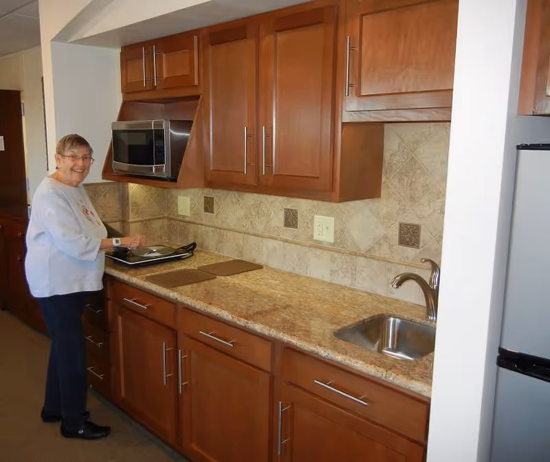 An elderly woman standing in a kitchen area with wooden cabinets, a microwave, a small sink, and a countertop. She is smiling and appears to be preparing food on a tray.