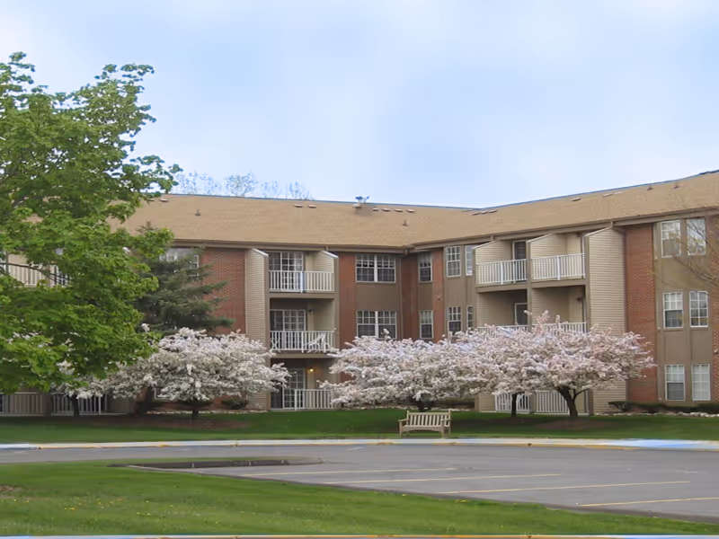 Three-story brick and siding senior living building with balconies, flowering trees, a bench, and a parking area in front.