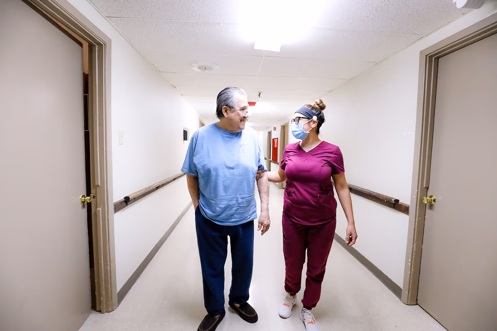 A healthcare worker in maroon scrubs and a face mask is walking down a hallway with an elderly man wearing a light blue shirt and dark pants. They are smiling and the healthcare worker has her hand gently on the man's arm. The hallway has white walls, beige doors, and handrails on both sides.