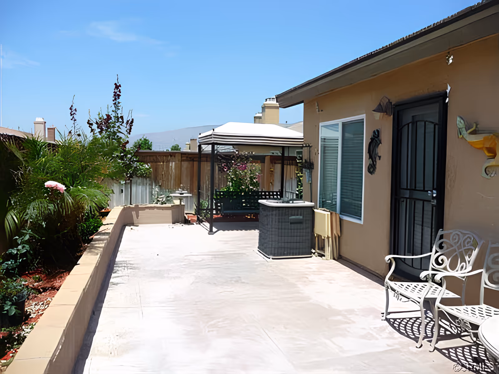 Sunlit fenced backyard patio with a gazebo swing, plants along a raised planter, and metal chairs by the house door.