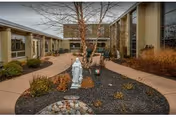 Outdoor courtyard area at a senior living facility with a statue of a religious figure surrounded by landscaping, including a small rock garden, bushes, and a tree. The courtyard is enclosed by a building with large windows and a paved walkway.