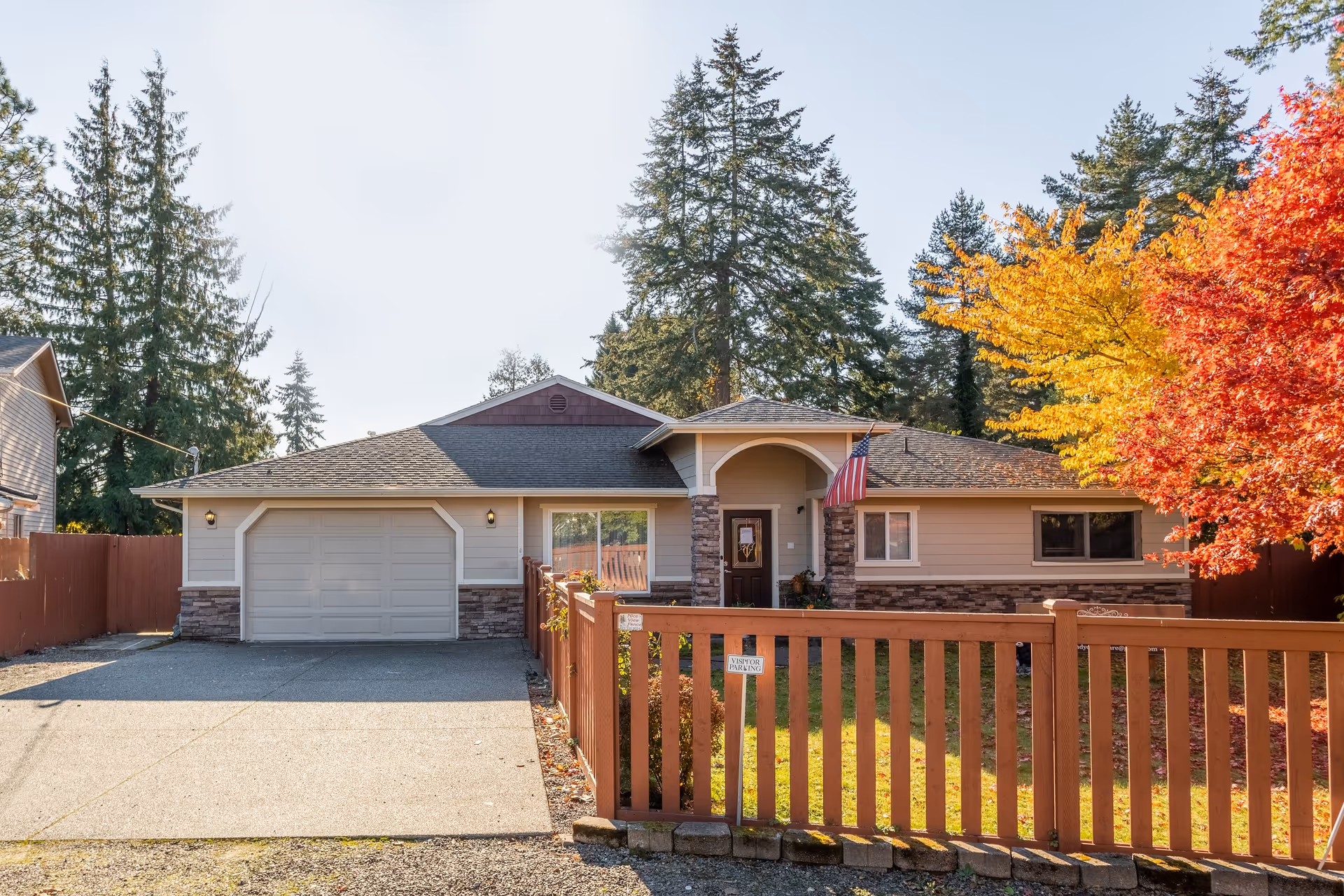 Front exterior view of a single-story house with a two-car garage, a wooden fence, and an American flag near the entrance. The yard has green grass and colorful autumn trees with red and yellow leaves. Tall evergreen trees are visible in the background under a clear sky.