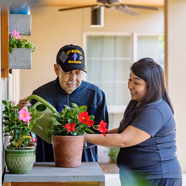 An elderly man wearing a 1st Marine Division cap waters potted red flowers on a table while a smiling woman assists him. They are outdoors near a building with windows and a ceiling fan visible in the background.