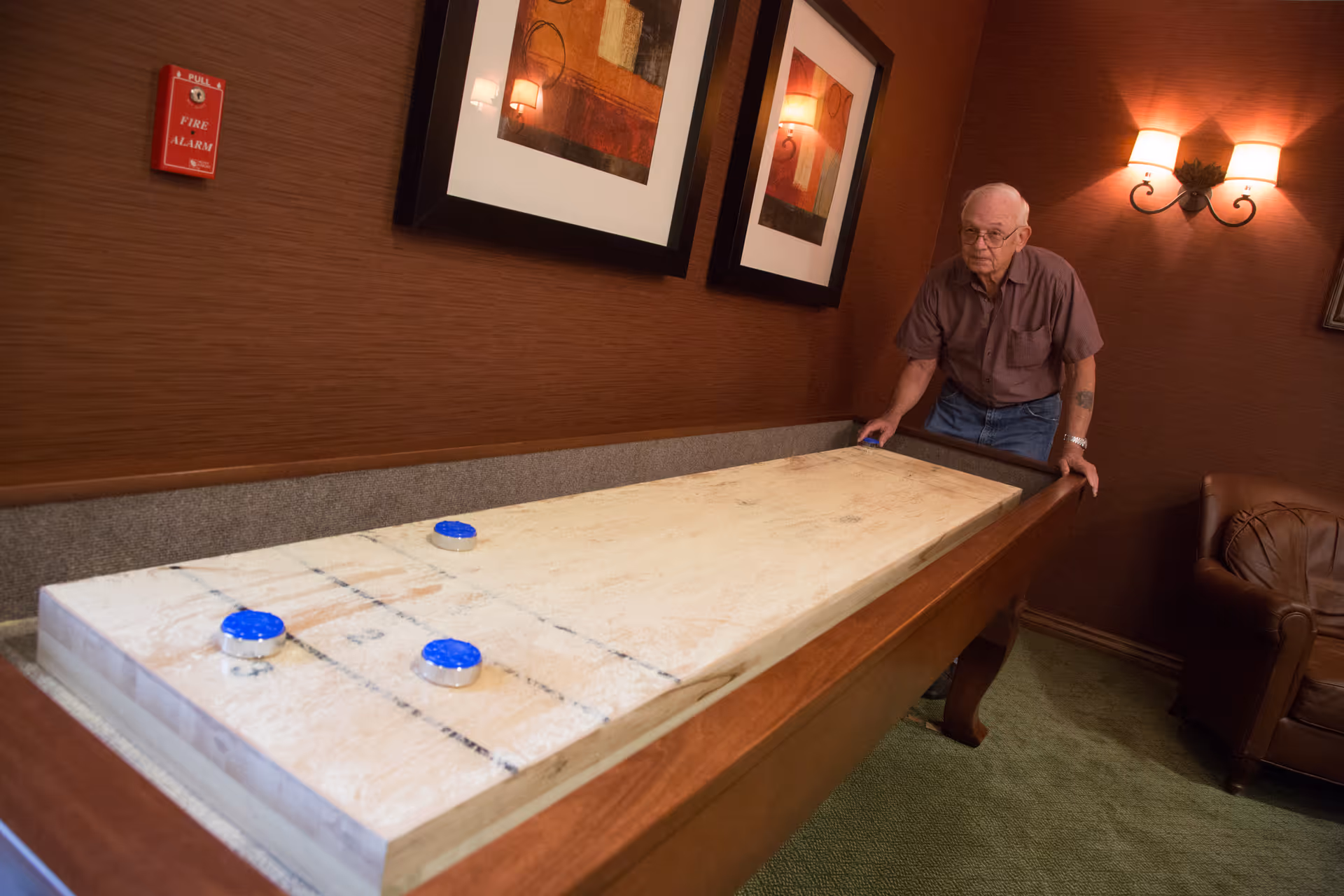 An elderly man playing shuffleboard indoors on a wooden shuffleboard table with three blue pucks on the board. The room has brown walls with two framed pictures, a fire alarm on the wall, a wall sconce light fixture, and a brown leather chair in the corner.