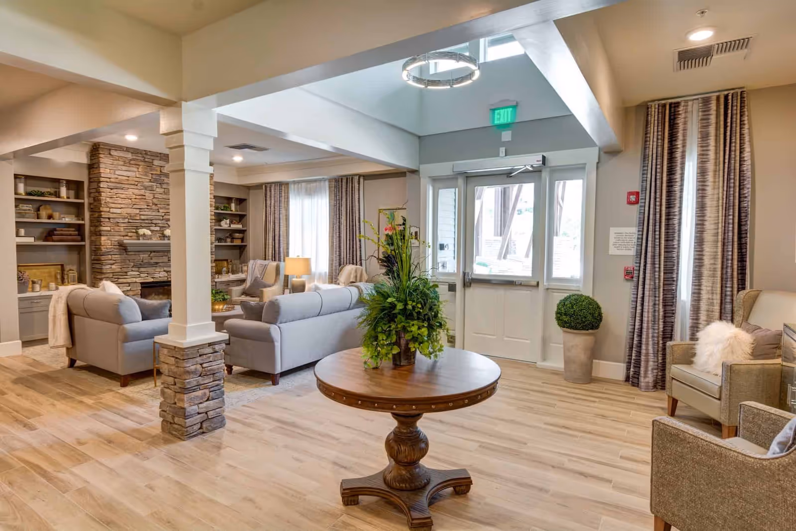 Bright, furnished communal living room with sofas and armchairs, a round center table with a plant, a stone fireplace and entry doors.