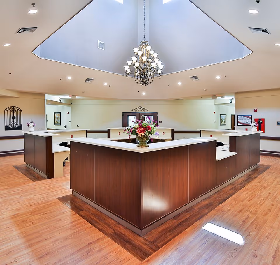 Interior view of a reception or nurse station area with a large wooden counter arranged in a U-shape, a floral arrangement on the counter, a chandelier hanging from a skylight ceiling, and wooden flooring.