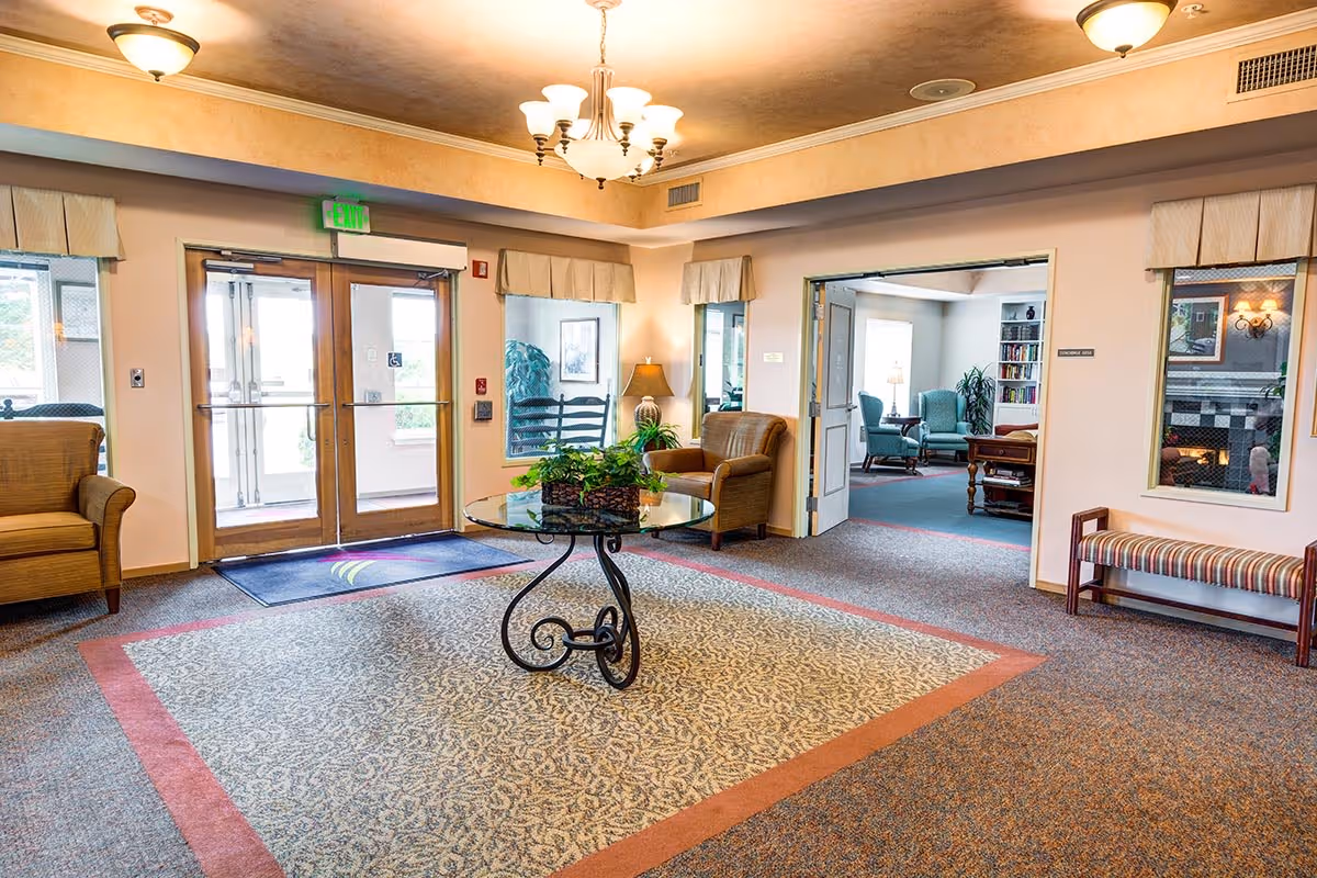 A bright and welcoming lobby area of a senior living facility with a glass-top round table holding a plant in the center. There are comfortable armchairs and a bench along the walls, with large windows and double glass doors leading outside. The room has warm lighting from ceiling fixtures and a chandelier, with a carpeted floor featuring a patterned design. Through an open doorway, a cozy sitting room with armchairs, a bookshelf, and a fireplace is visible.