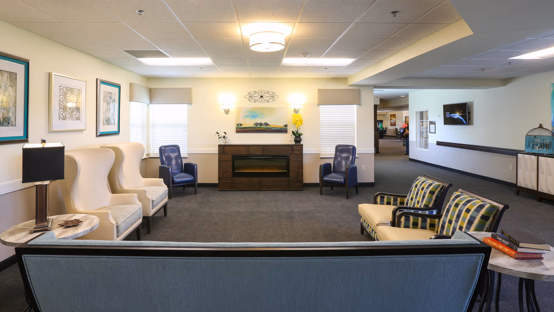 A cozy senior living facility common area with various seating options including beige wingback chairs, blue armchairs, and patterned cushioned chairs arranged around a fireplace. The room is softly lit with wall sconces and ceiling lights, decorated with framed artwork and a small table with books and a lamp.