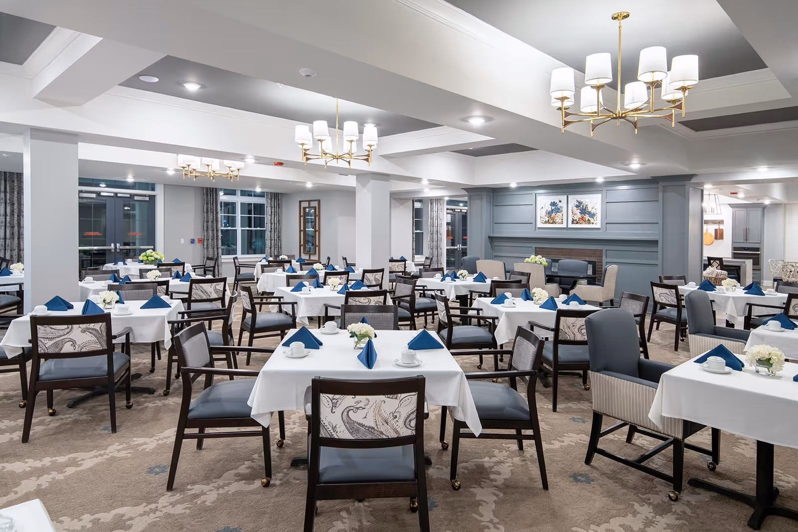 A spacious dining room in a senior living facility with multiple tables covered in white tablecloths, each set with blue folded napkins, white cups, and small floral centerpieces. The room features comfortable chairs, chandeliers, large windows with curtains, and a decorative fireplace with artwork above it.