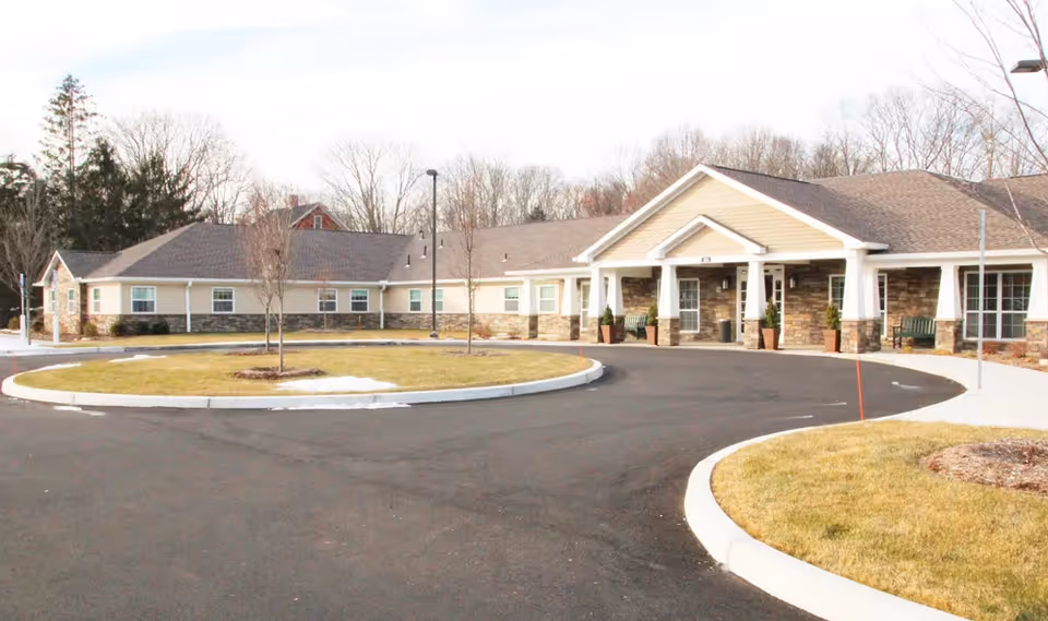 Exterior view of a single-story senior living facility building with a circular driveway and landscaped grassy areas with small trees. The building has a beige and stone facade with a covered entrance supported by white columns.