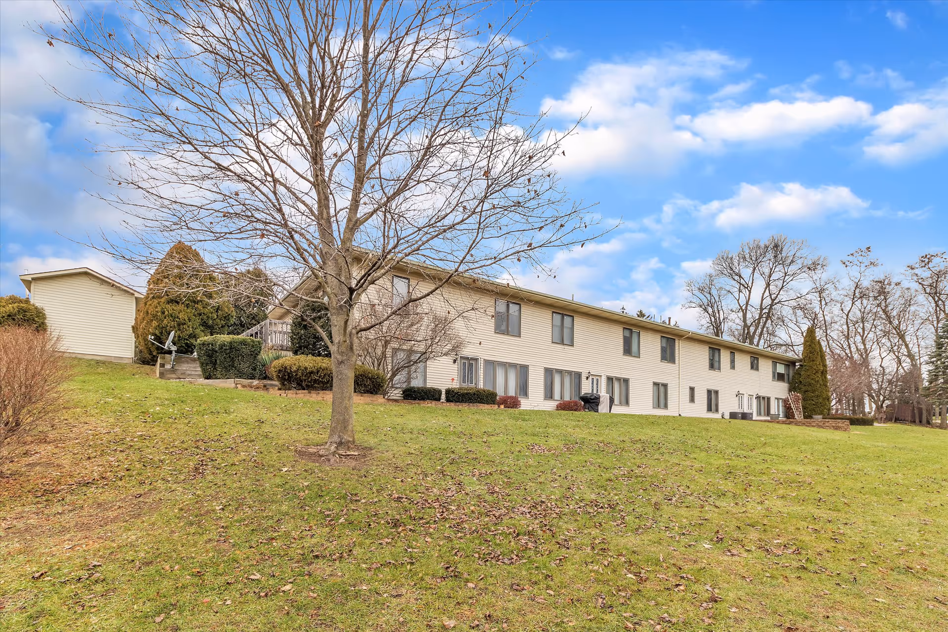Two-story beige apartment-style building on a grassy lawn with a leafless tree in the foreground under a partly cloudy sky.