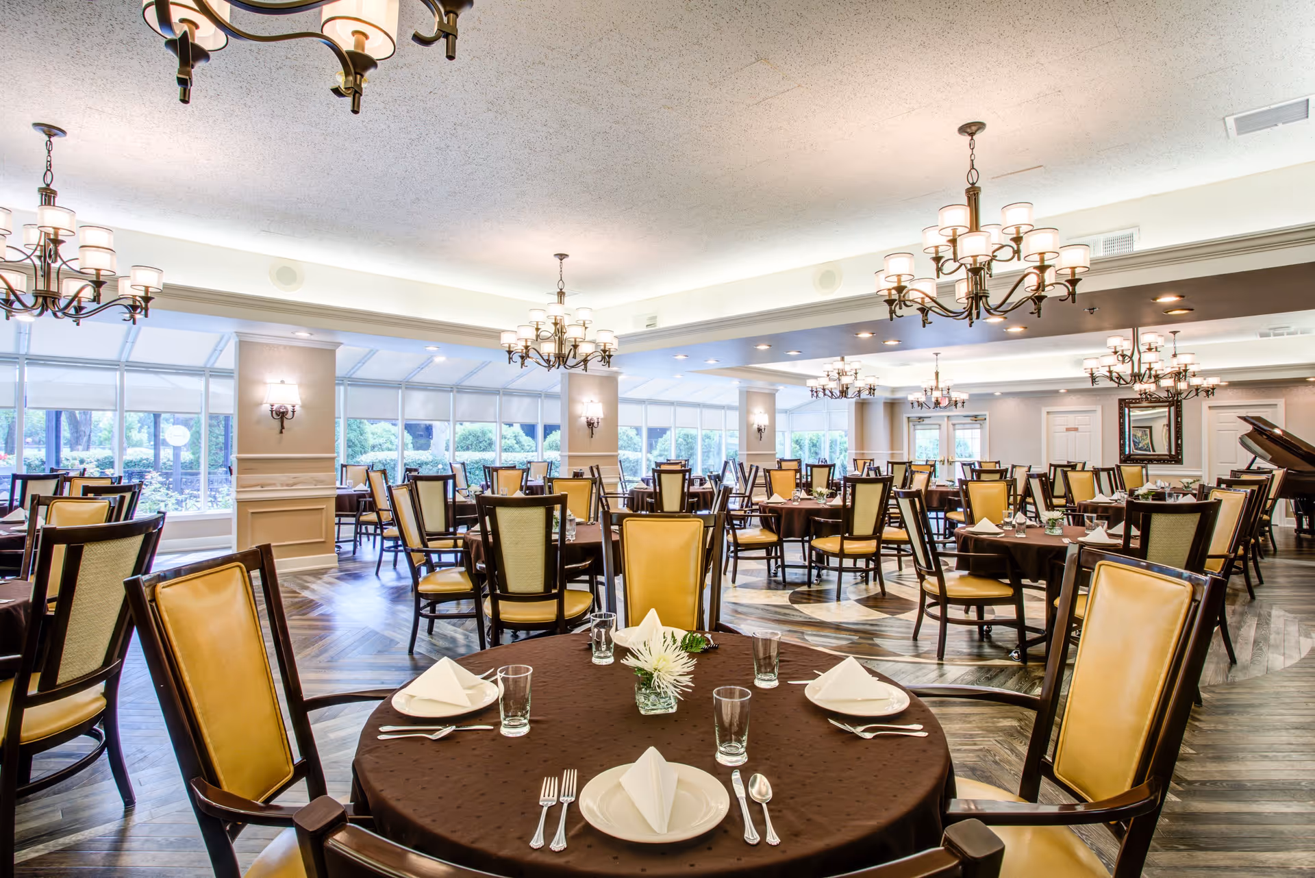 A spacious and elegant dining room with multiple round tables covered in brown tablecloths, each set with white plates, folded napkins, glasses, and silverware. The chairs have wooden frames with yellow cushioned backs and seats. Large windows line the walls, allowing natural light to fill the room. Several chandeliers hang from the ceiling, providing warm lighting. A grand piano is visible in the background near the wall with a mirror.