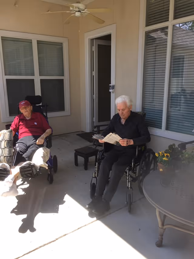 Two elderly men sitting outside on a covered patio area. One man is wearing a red shirt and cap, sitting in a wheelchair with his legs elevated. The other man, dressed in black, is also in a wheelchair and is reading a newspaper. There is a round glass table with a flower pot on it to the right, and windows and a door behind them.