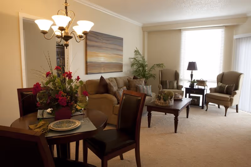 A cozy living room and dining area with a round dining table set for two, a beige sofa with multiple cushions, two armchairs, a wooden coffee table with decorative items, a side table with a lamp, a large window with blinds, a chandelier above the dining table, and a large wall painting depicting a sunset.