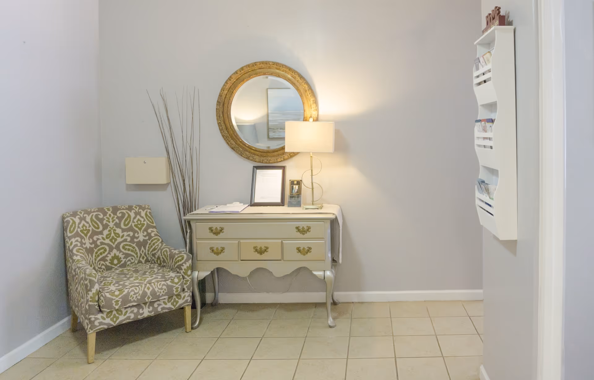 A small interior corner featuring a patterned armchair, a vintage-style cream-colored console table with drawers, a round gold-framed mirror above the table, a table lamp, and decorative tall sticks in a vase. There is also a white wall-mounted organizer on the right wall holding brochures or papers.