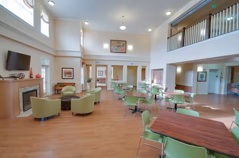 Spacious and well-lit common area in a senior living facility with wooden floors, green chairs around tables, a seating area with armchairs near a fireplace and a wall-mounted TV, and large windows letting in natural light.