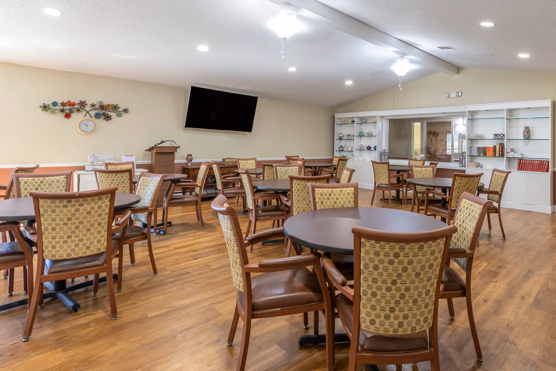 A spacious dining room with multiple round tables and wooden chairs with patterned upholstery. The room has a wooden floor, a wall-mounted flat screen TV, a decorative wall clock with floral accents, and built-in white shelves displaying various decorative items and books. The ceiling has recessed lighting and ceiling fans.
