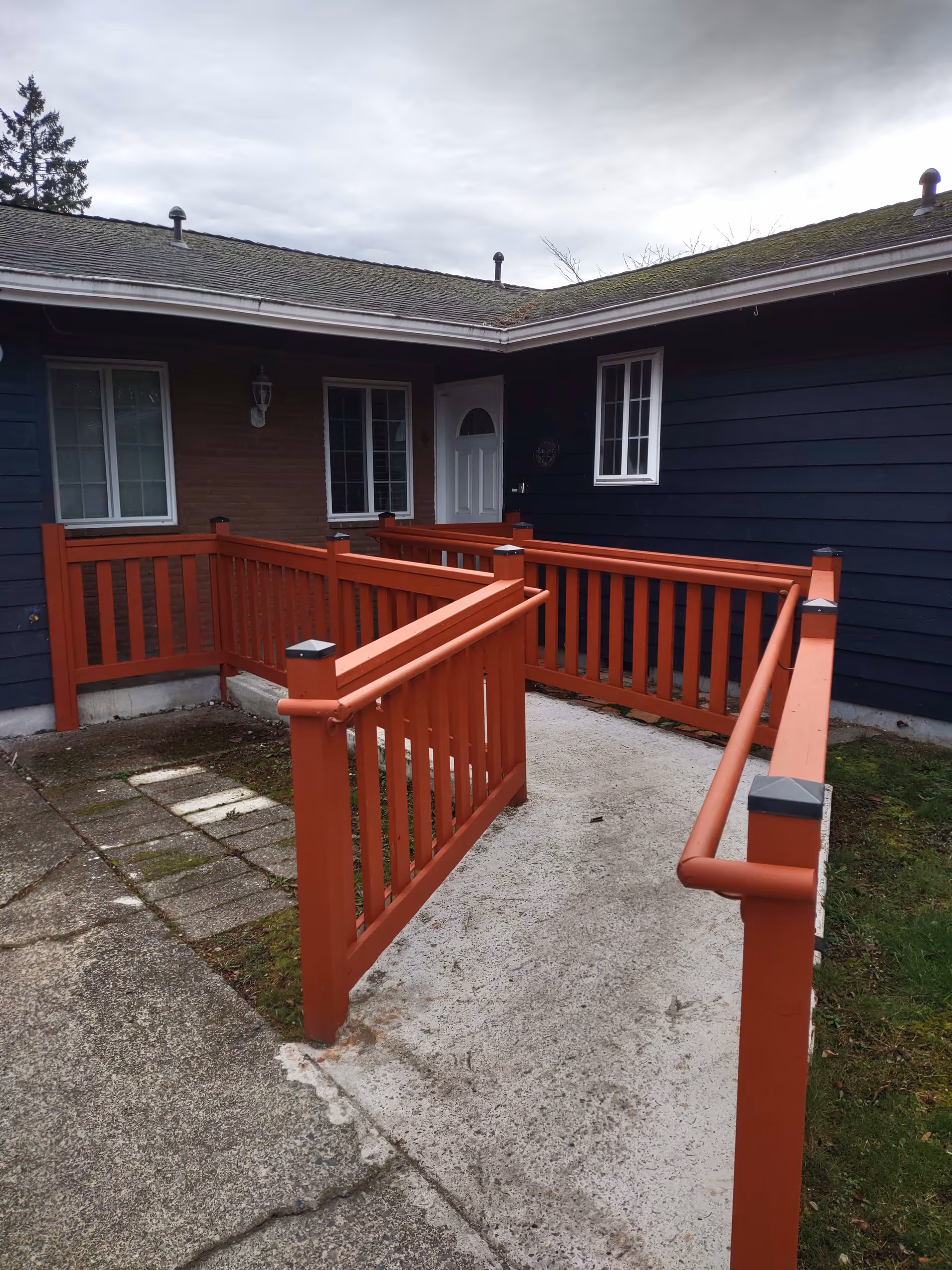 Exterior view of a building corner with a concrete wheelchair ramp featuring red wooden railings leading to a white door. The building has dark blue and brown siding with two windows visible. The sky is overcast.
