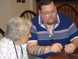 An elderly woman and a younger man sitting at a wooden table engaged in a hands-on activity. The man is showing the woman something with his hands, possibly involving string or yarn. Both are focused on the task. The setting appears to be indoors with wooden chairs in the background.
