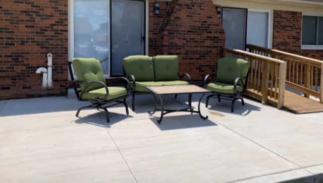 Outdoor patio area with three green cushioned chairs and a rectangular table on a concrete surface, adjacent to a brick building with windows and a wooden ramp.