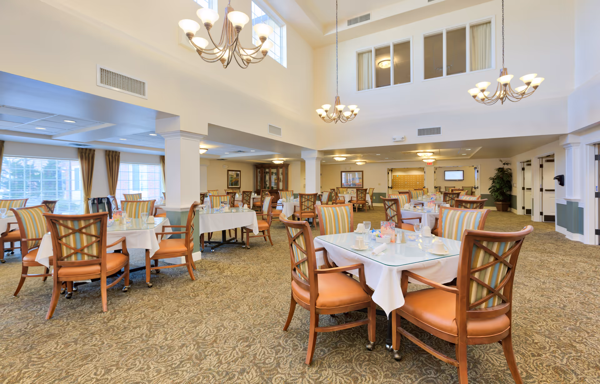 A spacious dining room with multiple tables covered in white tablecloths, each set with cups, glasses, and napkins. The chairs have wooden frames with striped upholstery. The room features high ceilings with chandeliers, large windows with curtains, and carpeted floors with a patterned design.