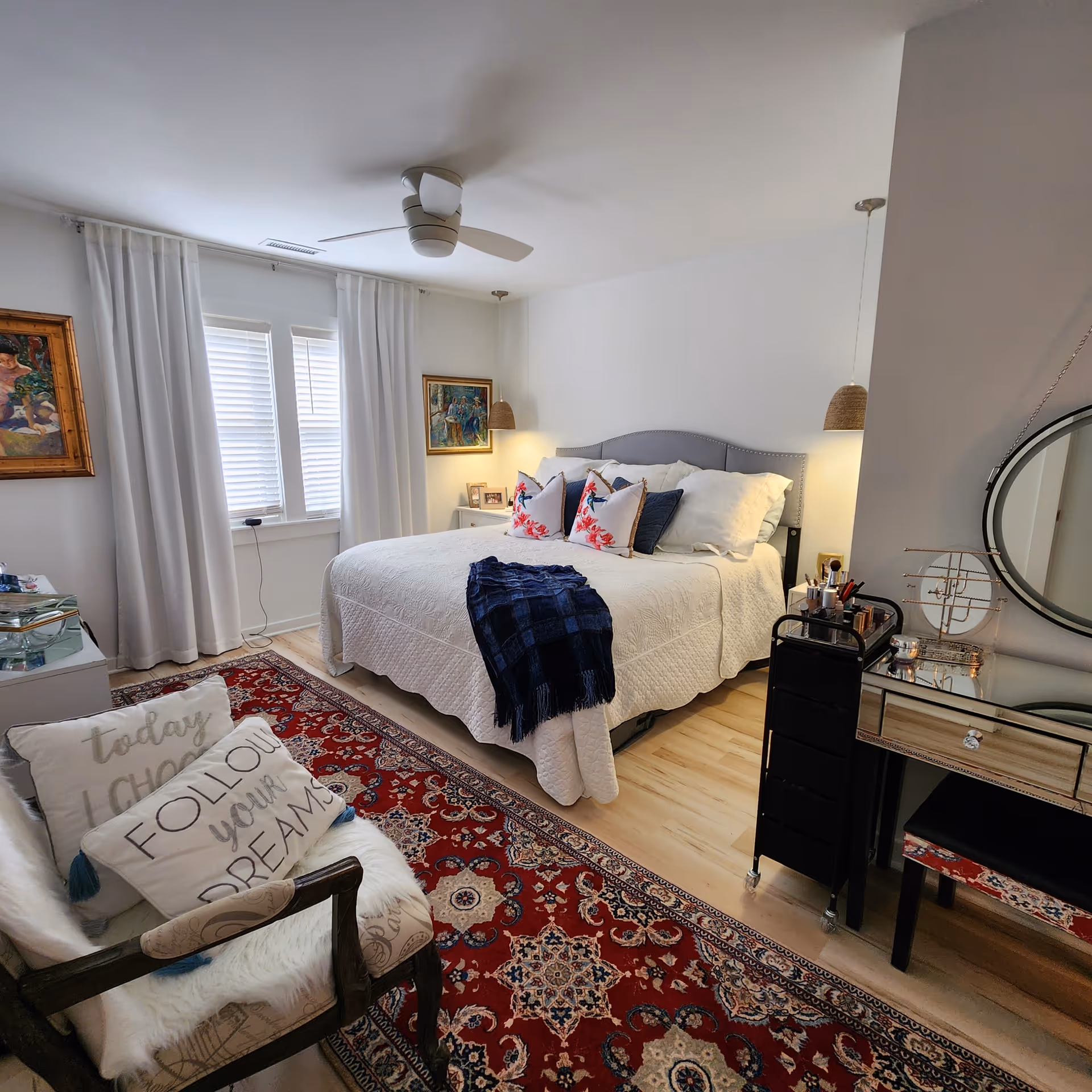 A cozy bedroom with a large bed covered in white bedding and decorative pillows, a blue throw blanket at the foot of the bed, a red patterned rug on the floor, a chair with cushions that say 'today I CHOOSE' and 'FOLLOW your DREAMS', two framed paintings on the walls, a ceiling fan, and a vanity table with a round mirror and makeup items.